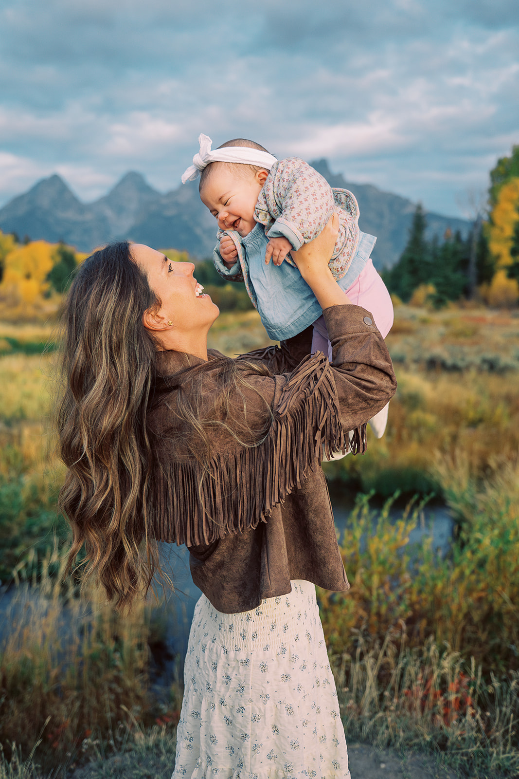 A happy mother in a brown jacket and tan dress plays with her giggling baby abov her head while standing by a river at sunset
