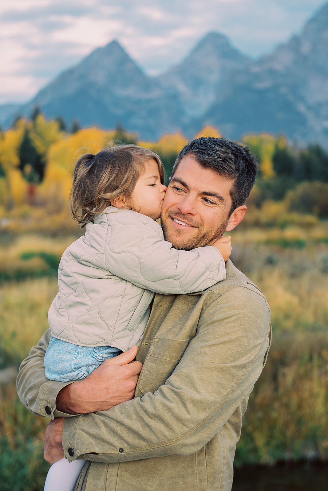 A toddler girl in a tan jacket kisses and hugs her dad while holding her on his hip while walking a meadow after visiting daycares in Jackson, WY
