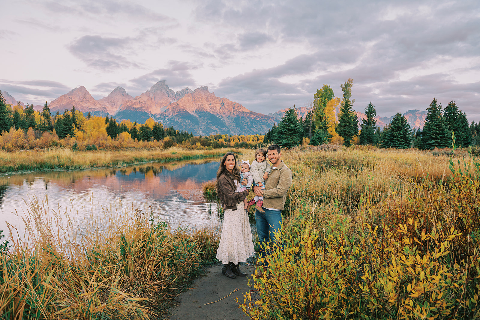 Happy mom and dad in brown and tan stand on a riverside trail at sunset holding their two toddlers on their hips after finding daycares in Jackson, WY