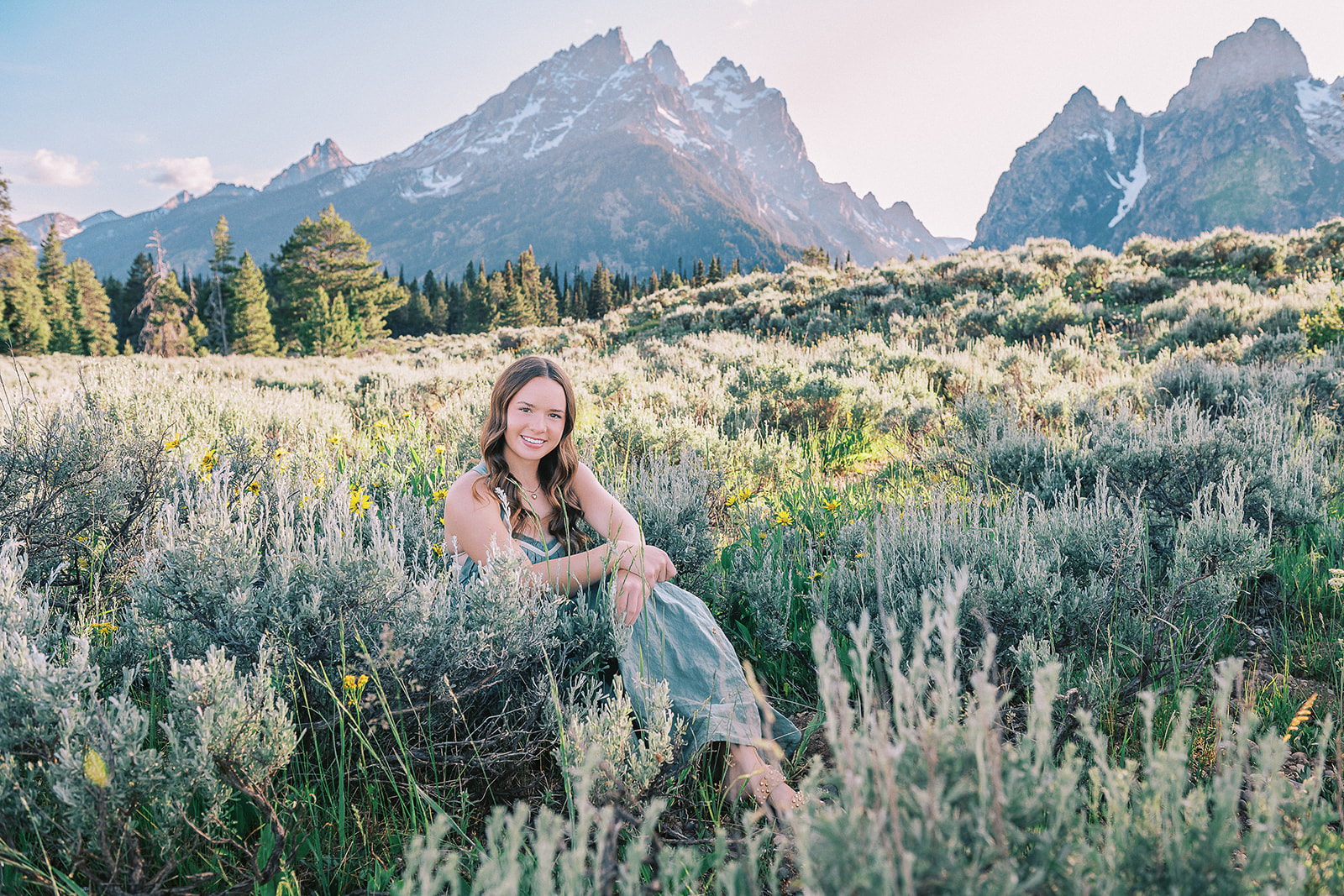 A high school senior in a green dress smiles while sitting in the grasses of a meadow at sunrise
