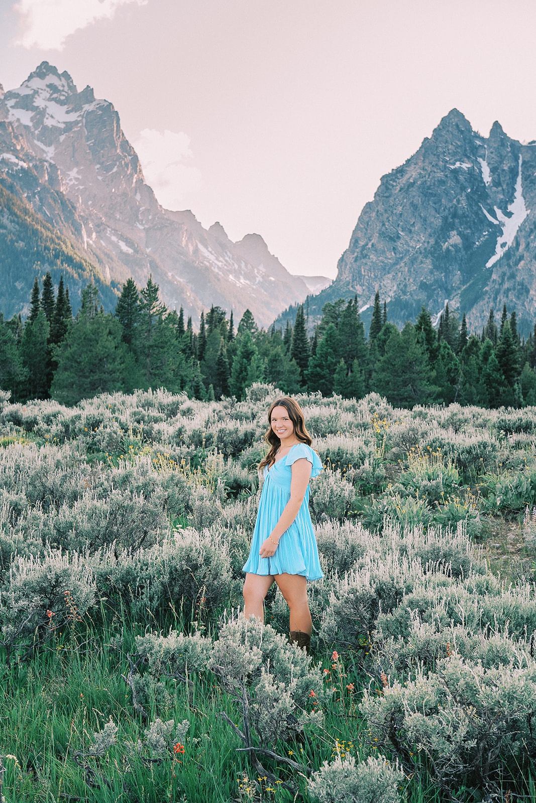 A high school senior in a blue dress walks in a meadow at sunrise in the mountains after visiting nail salons in Jackson, Wyoming