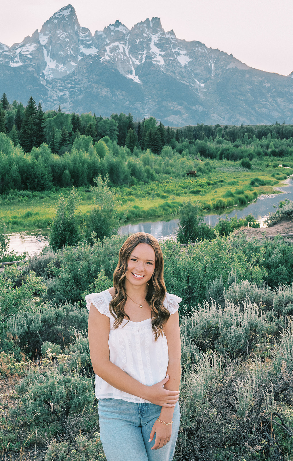 A high school senior stands smiling in a white blouse by a river under the tetons after finding nail salons in Jackson, Wyoming