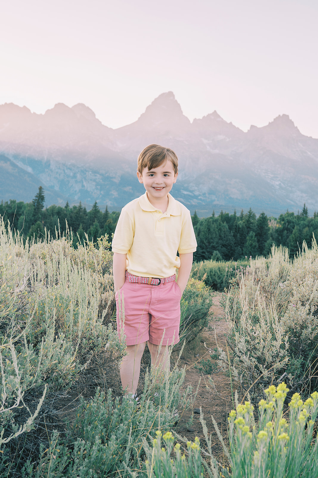 A smiling young boy in yellow and pink stands in a narrow trail in the mountains after attending private schools in Jackson, WY