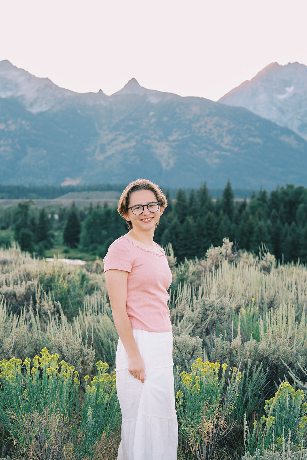 A high school senior in a pink shirt smiles while walking a trail of tall grasses at sunset after attending private schools in Jackson, WY