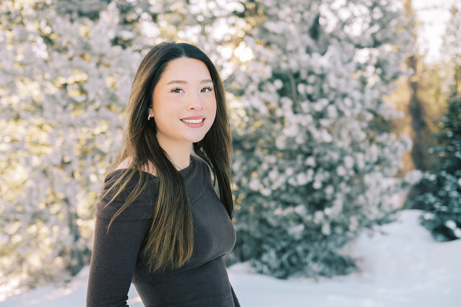 A teenage woman in a dark blouse smiles while walking in a snow covered forest at sunrise