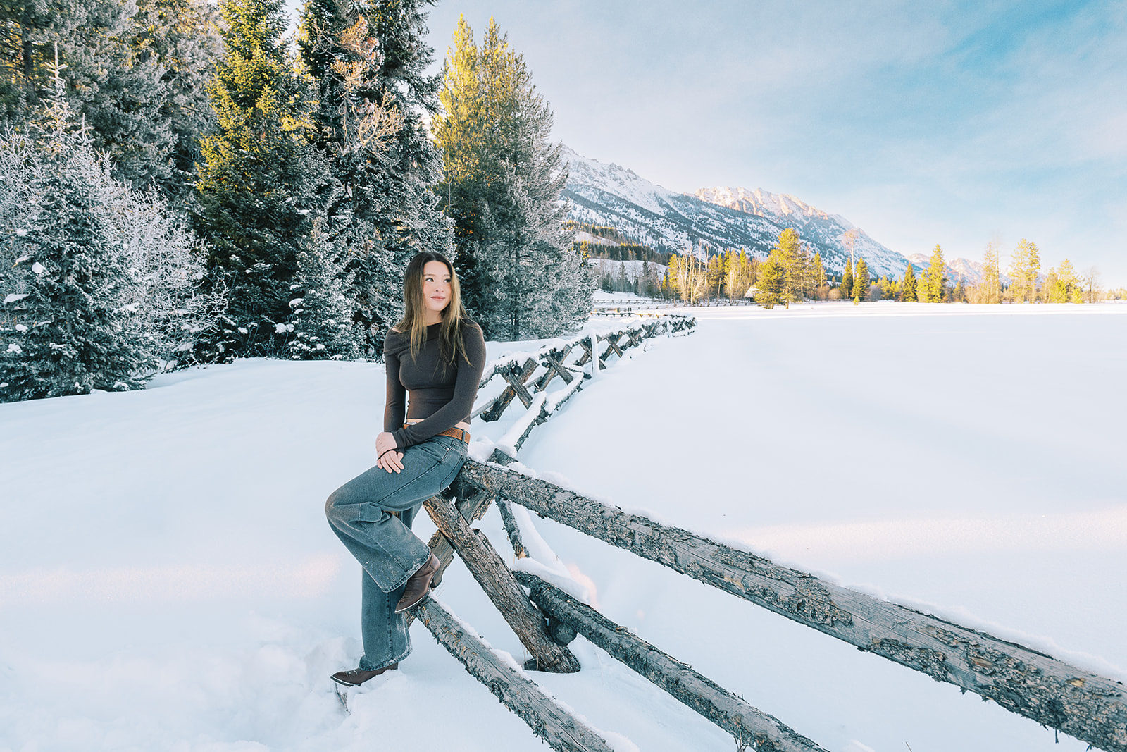 A high school senior who used test prep in Jackson, WY smiles while sitting on a pasture fence at sunrise in a dark top and jeans in the snow
