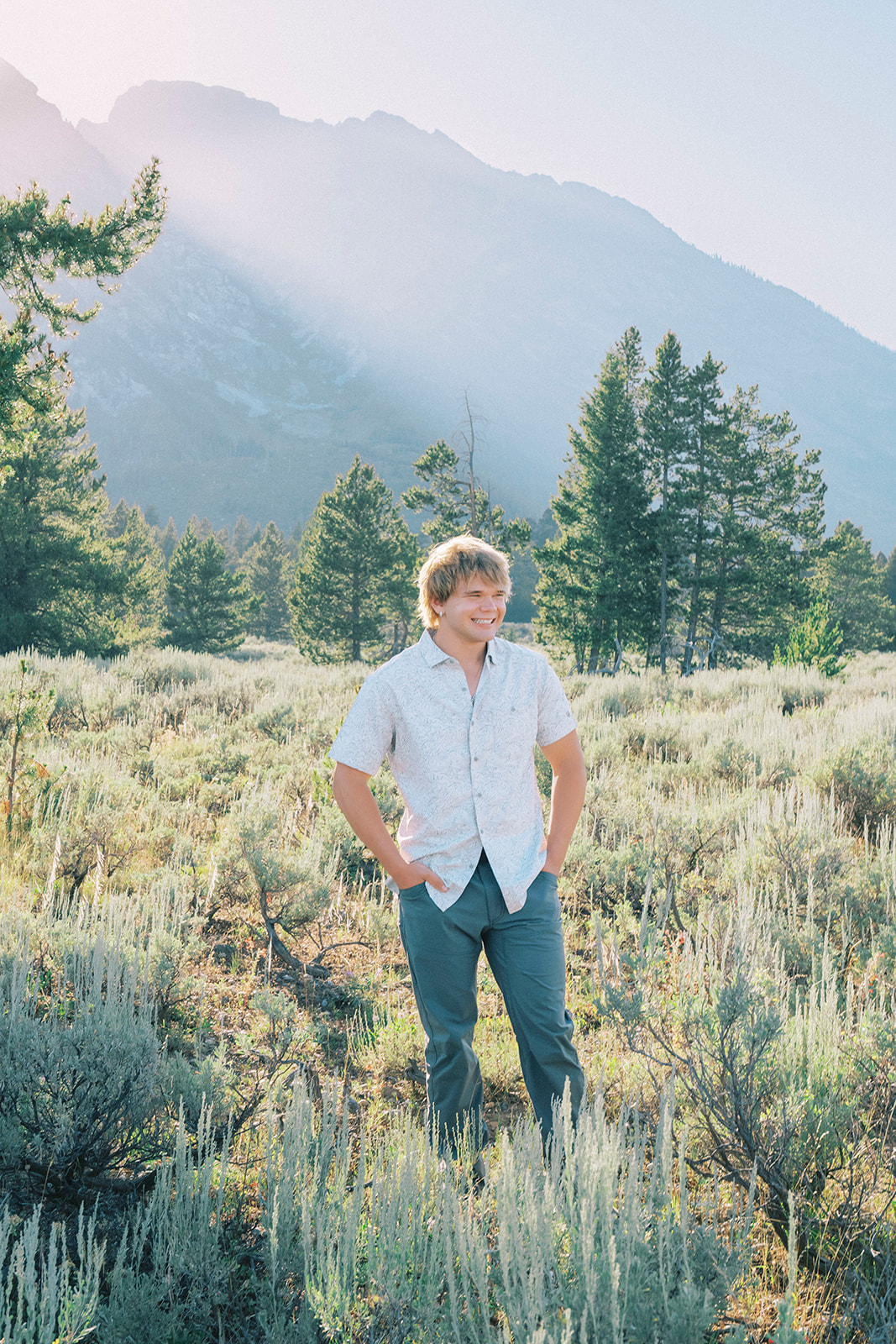 A smiling high school senior in a white shirt stands with hands in pockets at sunrise while on a hike near the mountains