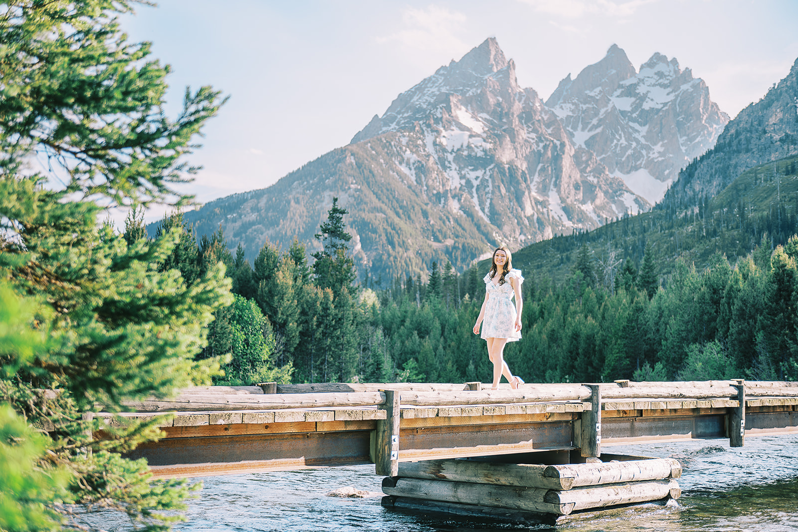 A smiling woman walks on a wooden bridge over a river in the mountains