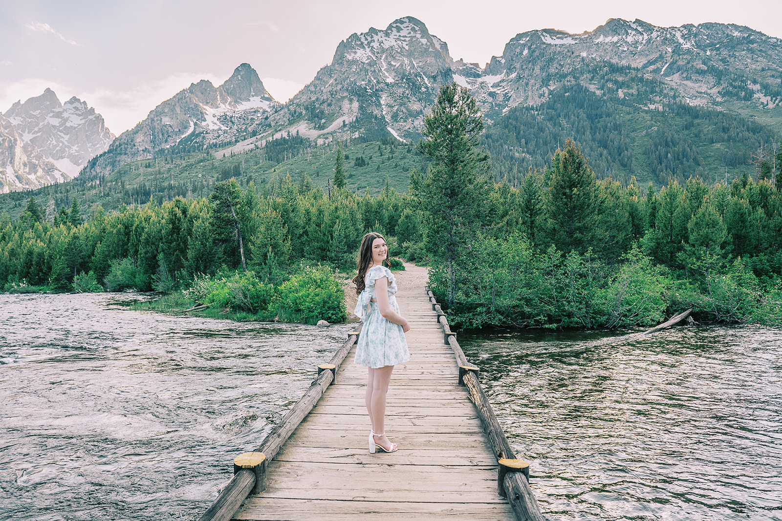 A high school senior in a blue and white dress walks over a wooden footbridge on a river smiling over her shoulder after finding hair salons in Jackson, WY