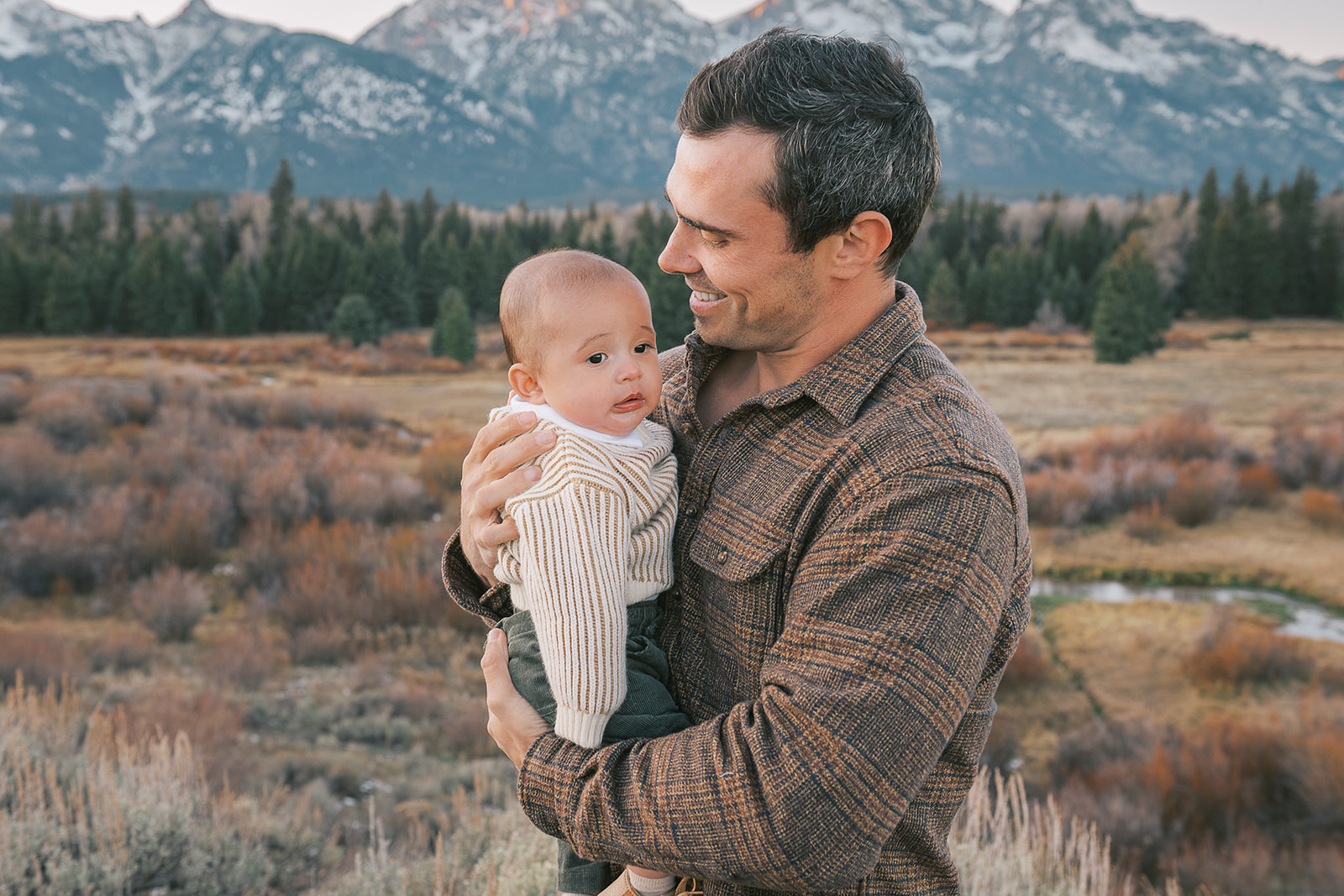 A happy dad smiles while cradling his baby in a plaid shirt in a meadow after finding Jackson Hole nannies