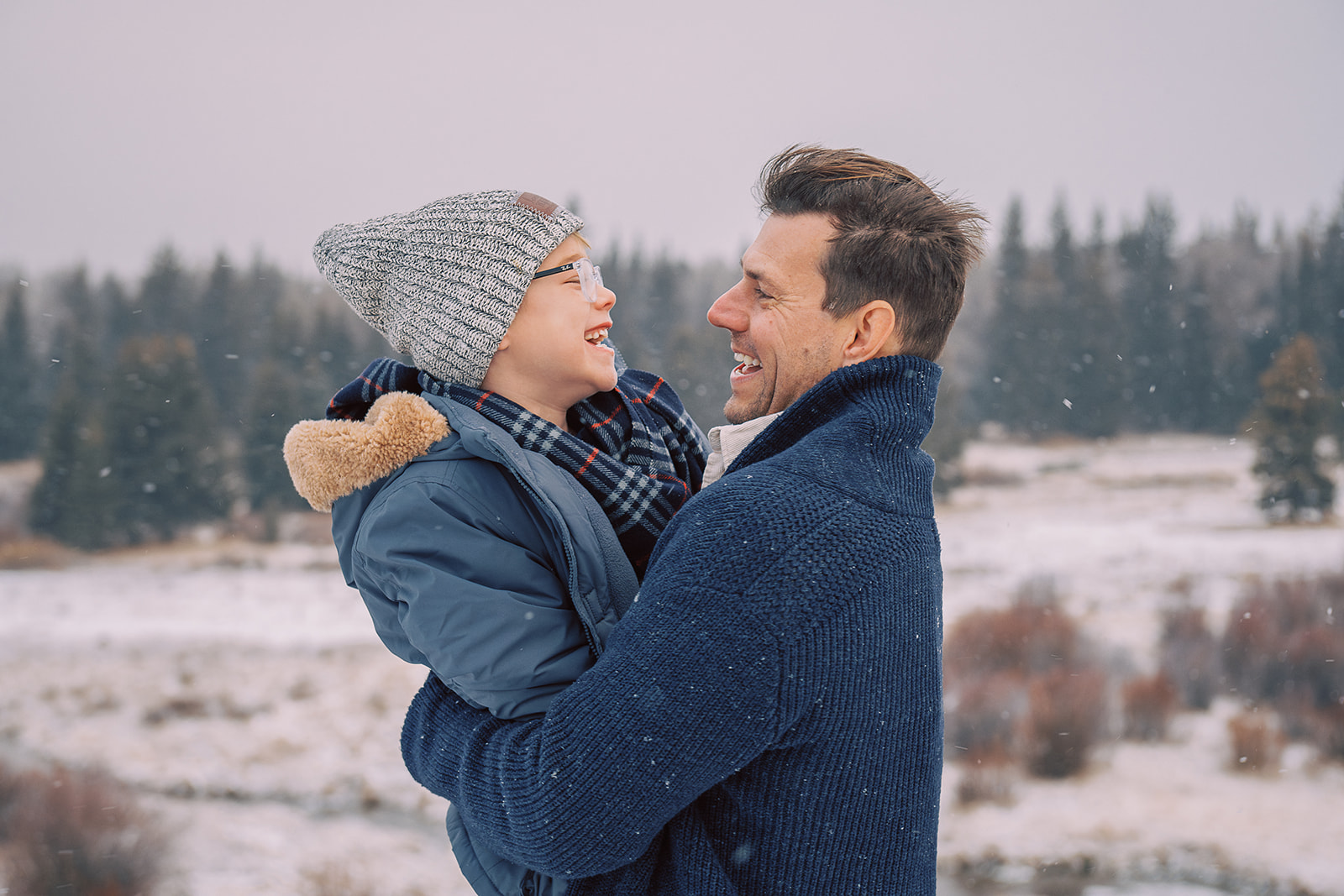 A happy dad in a blue sweater hugs and laughs with his toddler son as snow falls around them in a meadow before visiting toy store in Jackson, WY