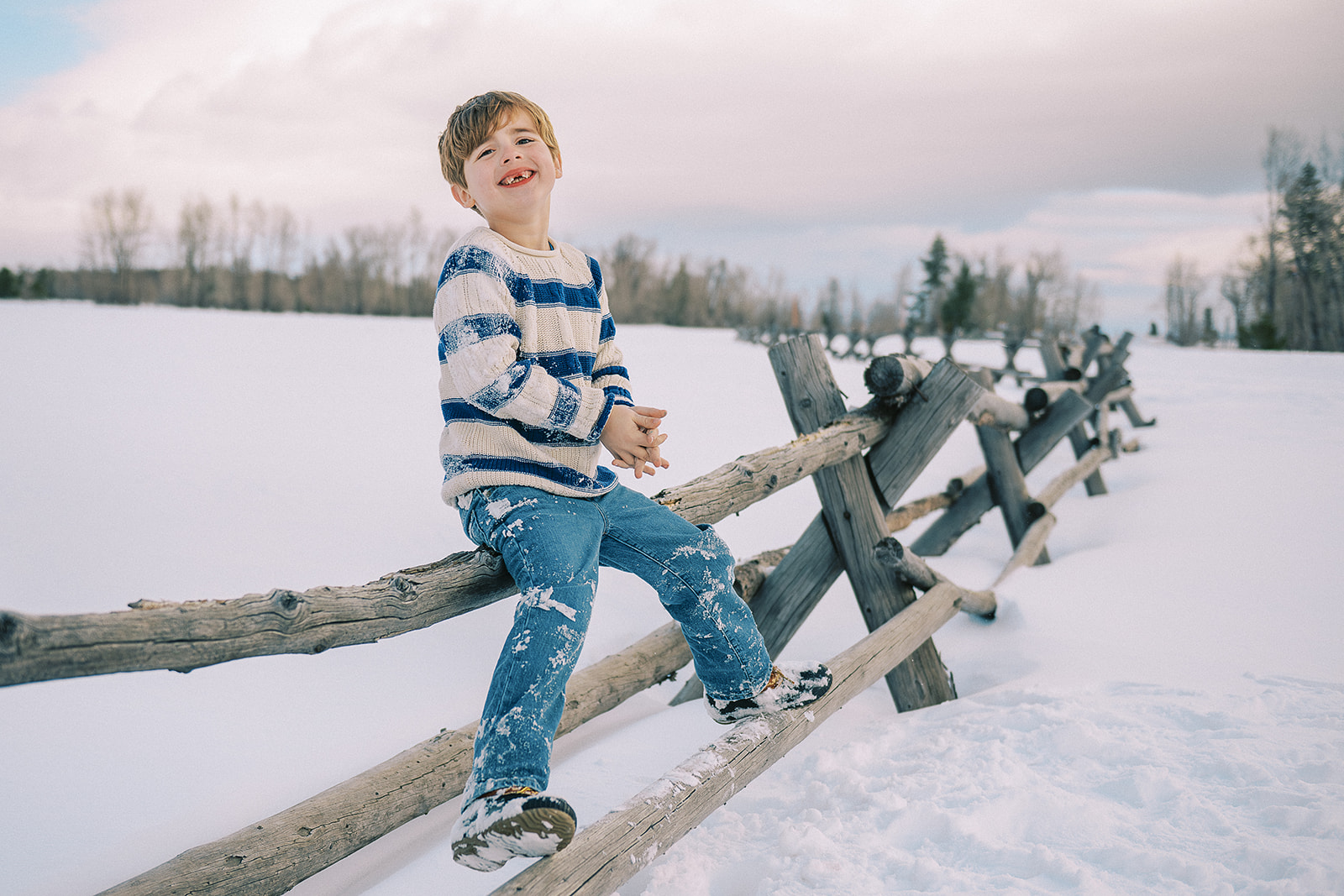 A giggling young boy with missing front teeth in a blue sweater covered in snow sits on a pasture fence after visiting pediatricians in Jackson, WY