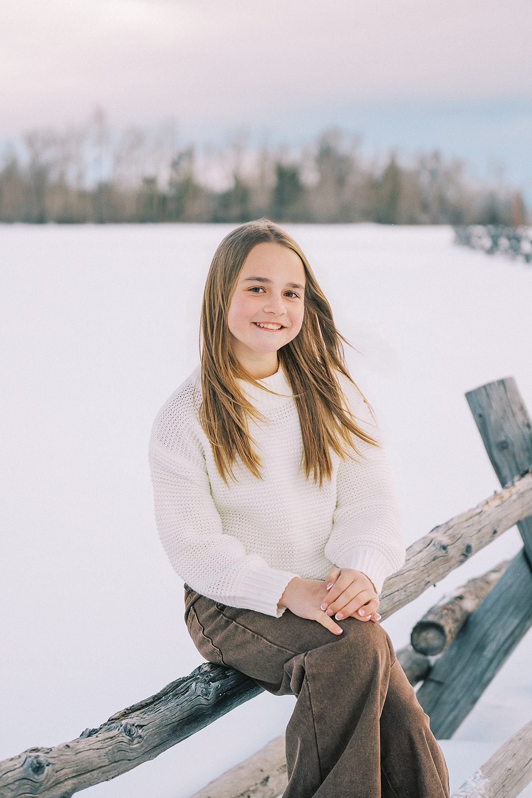 A happy young girl in a white sweater sits smiling on a rustic pasture fence after finding pediatricians in Jackson, WY