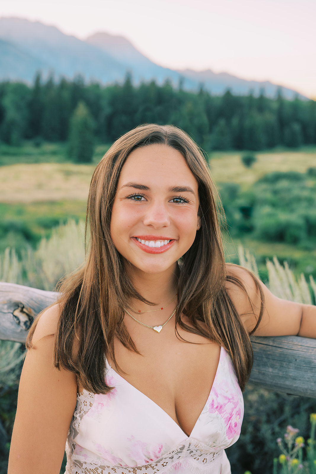 A closeup of a high school senior leaning on a rustic fence in a pink flower print dress at sunrise after finding volunteer opportunities in Jackson, WY