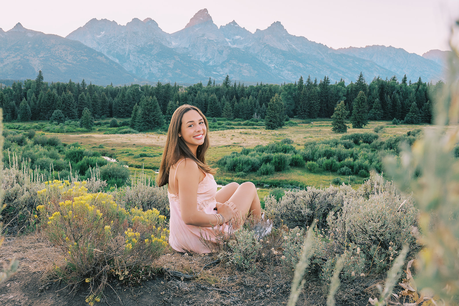 A woman in a pink dress sits on an overlook near the tetons smiling over her shoulder after enjoying volunteer opportunities in Jackson, WY