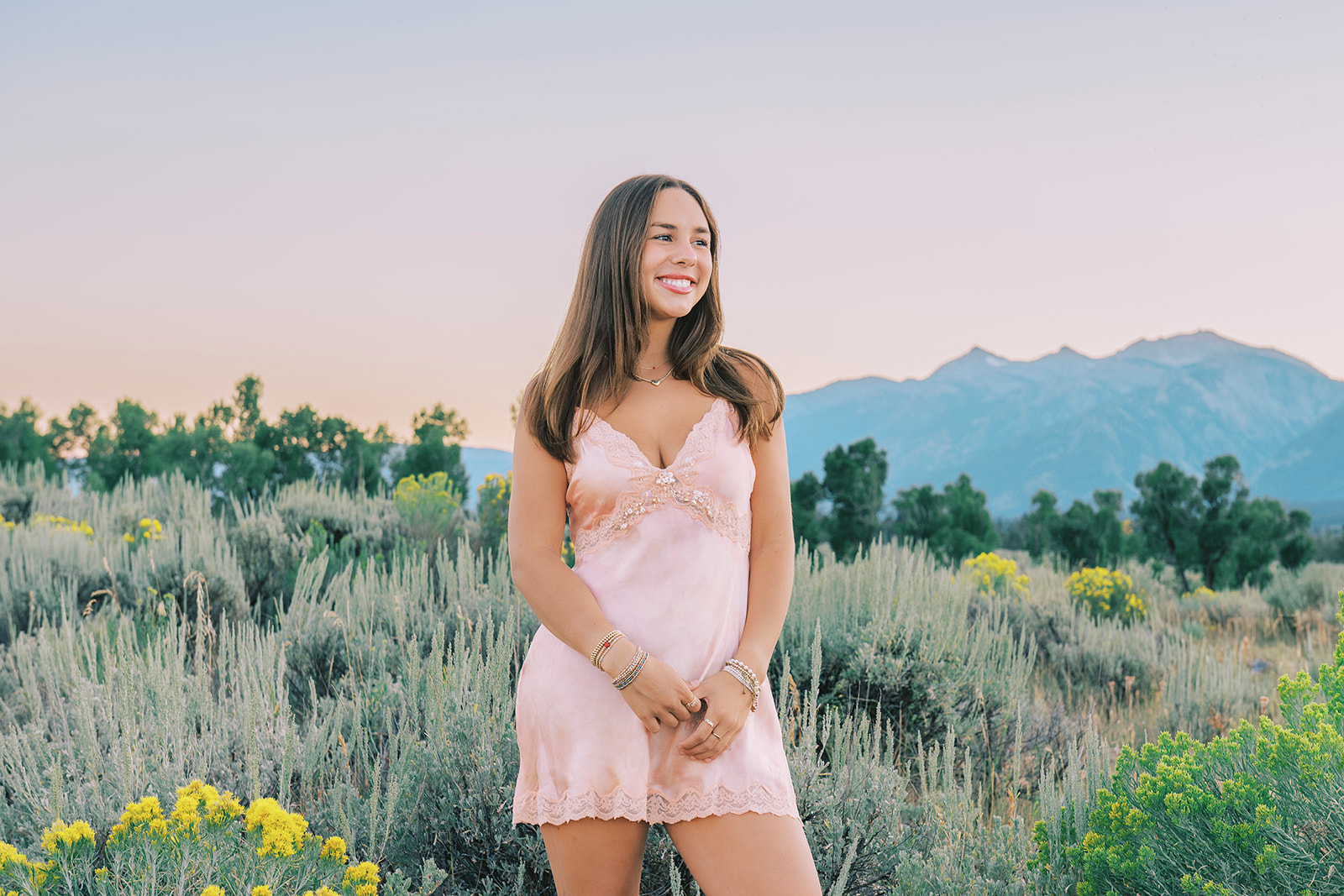 A high school senior in a pink dress stands smiling in a meadow filled with yellow flowers after finding volunteer opportunities in Jackson, WY