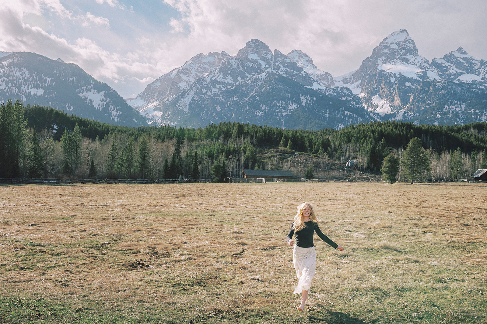 A laughing woman walks in a pasture in a black blouse and lace boho skirt barefoot near the mountains after visiting graduation party venues in Jackson, WY