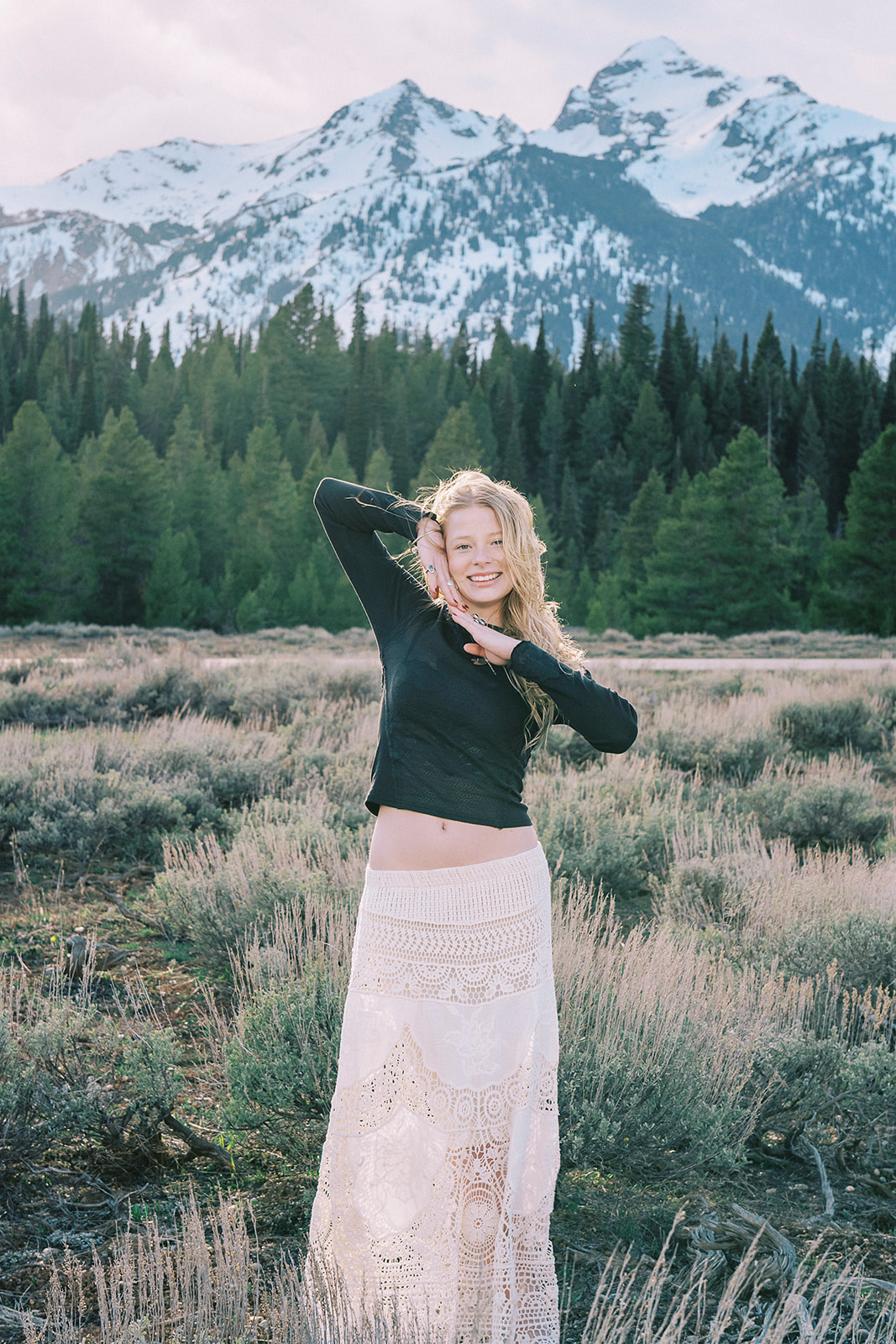 A happy high school senior in a black blouse and lace skirt stands in a valley below snow covered mountains with hands against her face