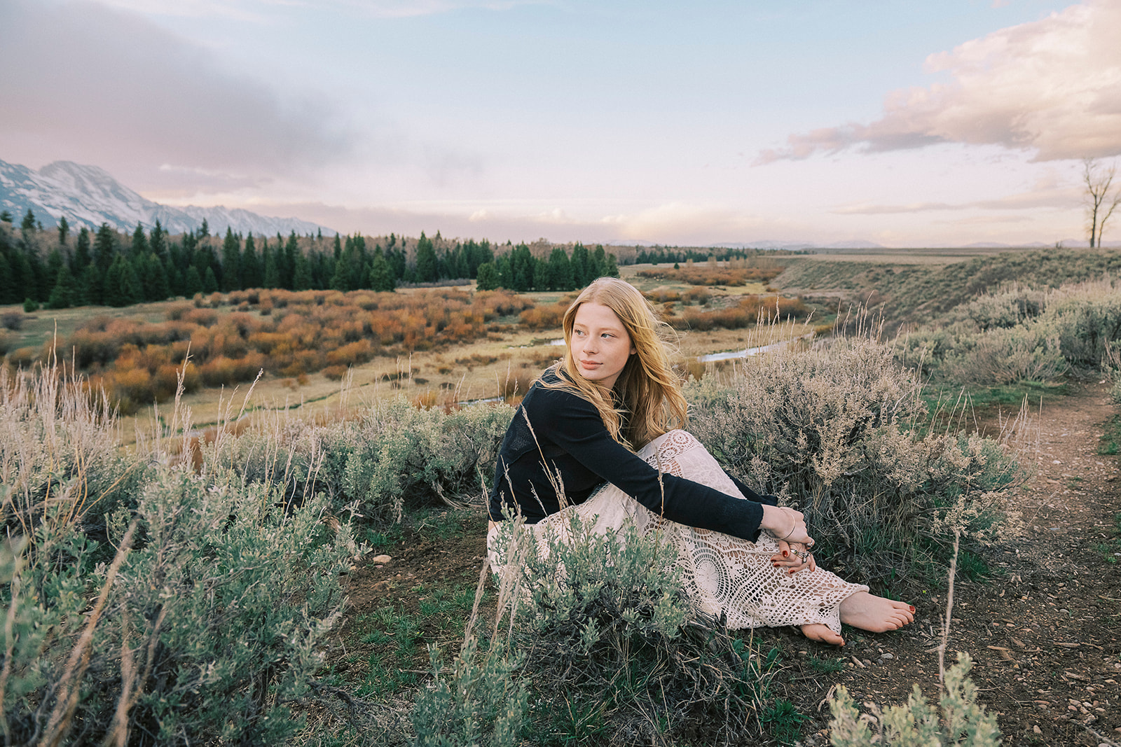 A high school grad in a lace skirt and black blouse sits on a hillside smiling over her shoulder at sunrise after exploring graduation party venues in Jackson, WY