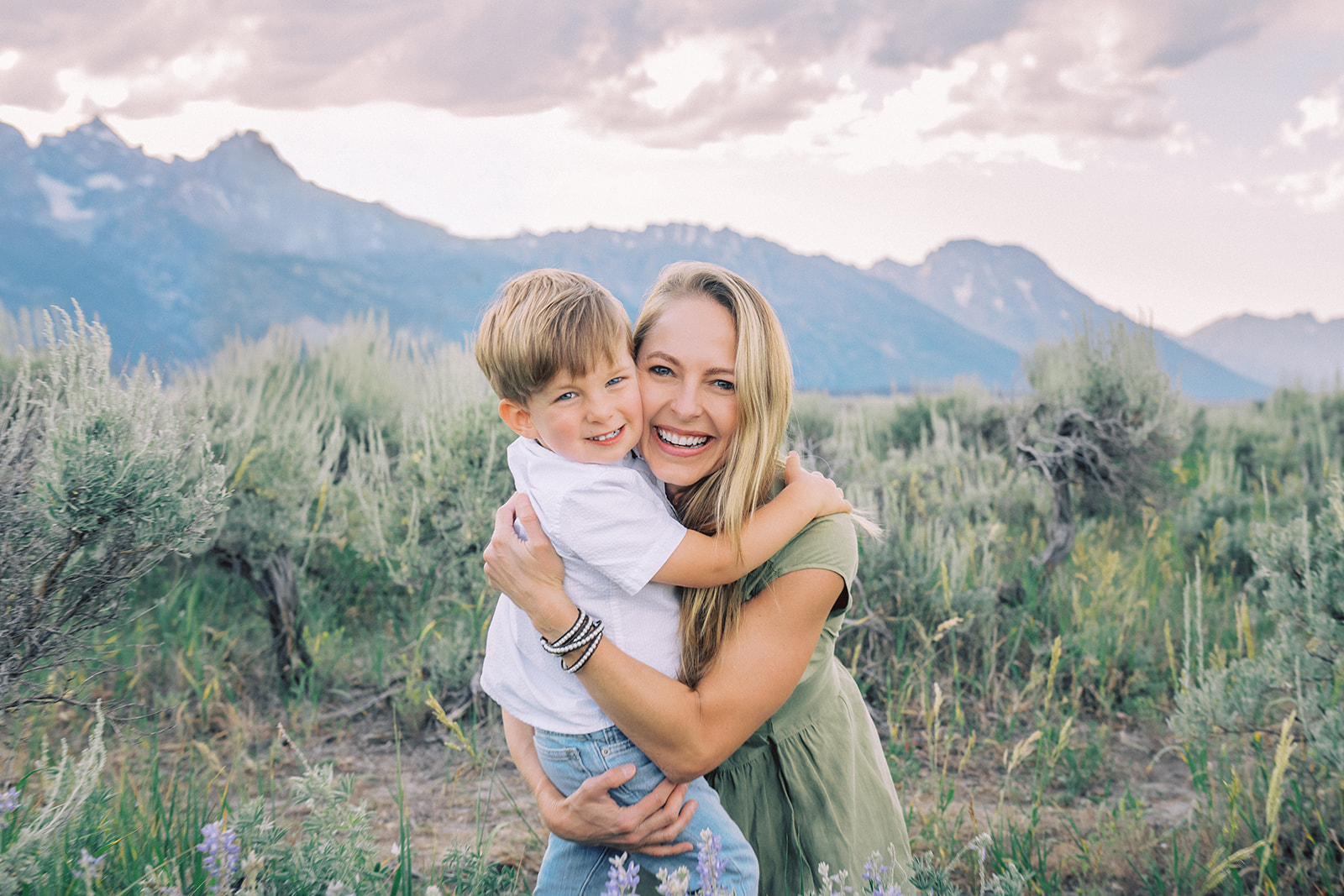 A happy mom in a green dress hugs her happy son in a white shirt in a valley below the tetons