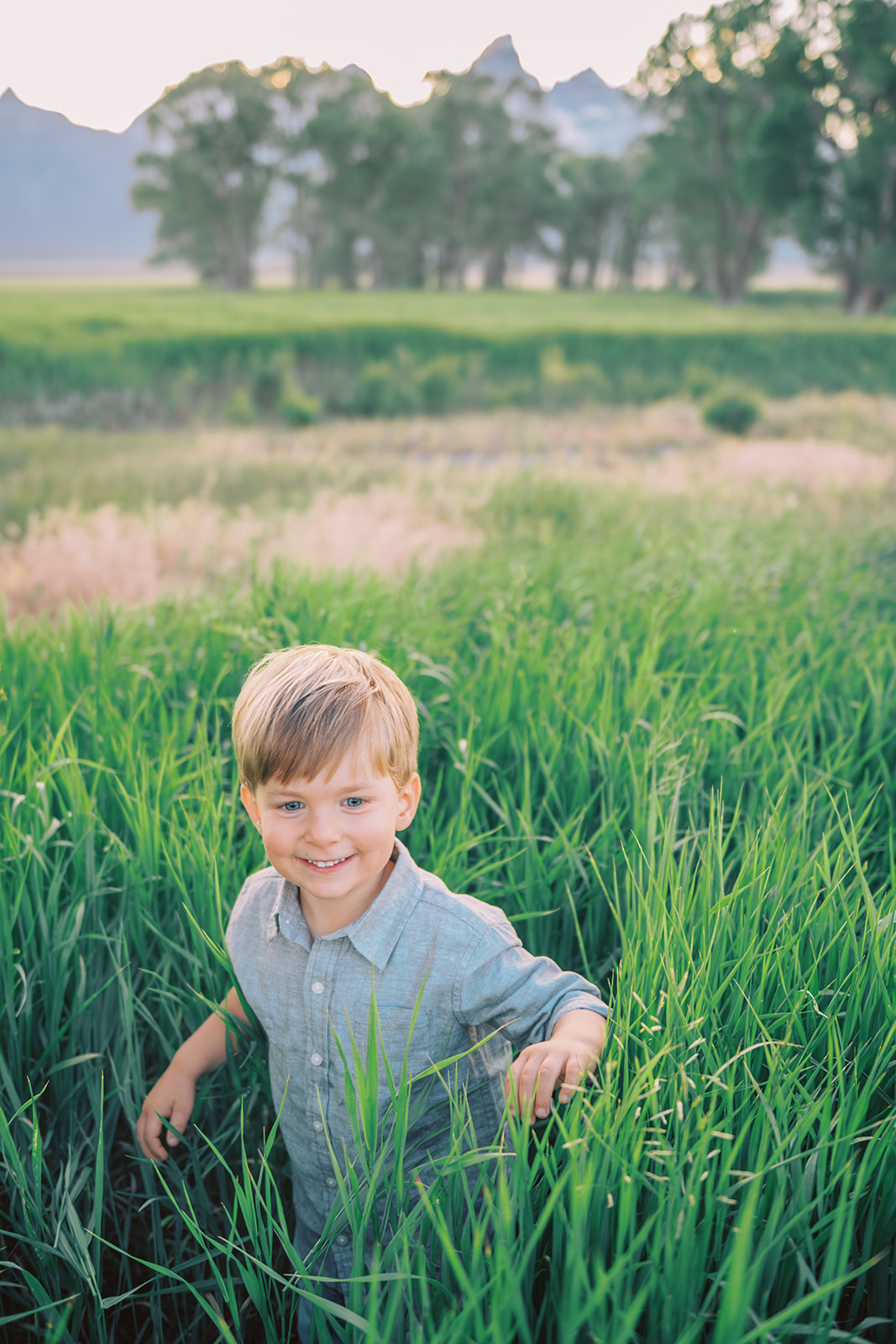A young boy in a grey button down walks through tall green grass with a big smile after enjoying pediatric dentists in Jackson, WY