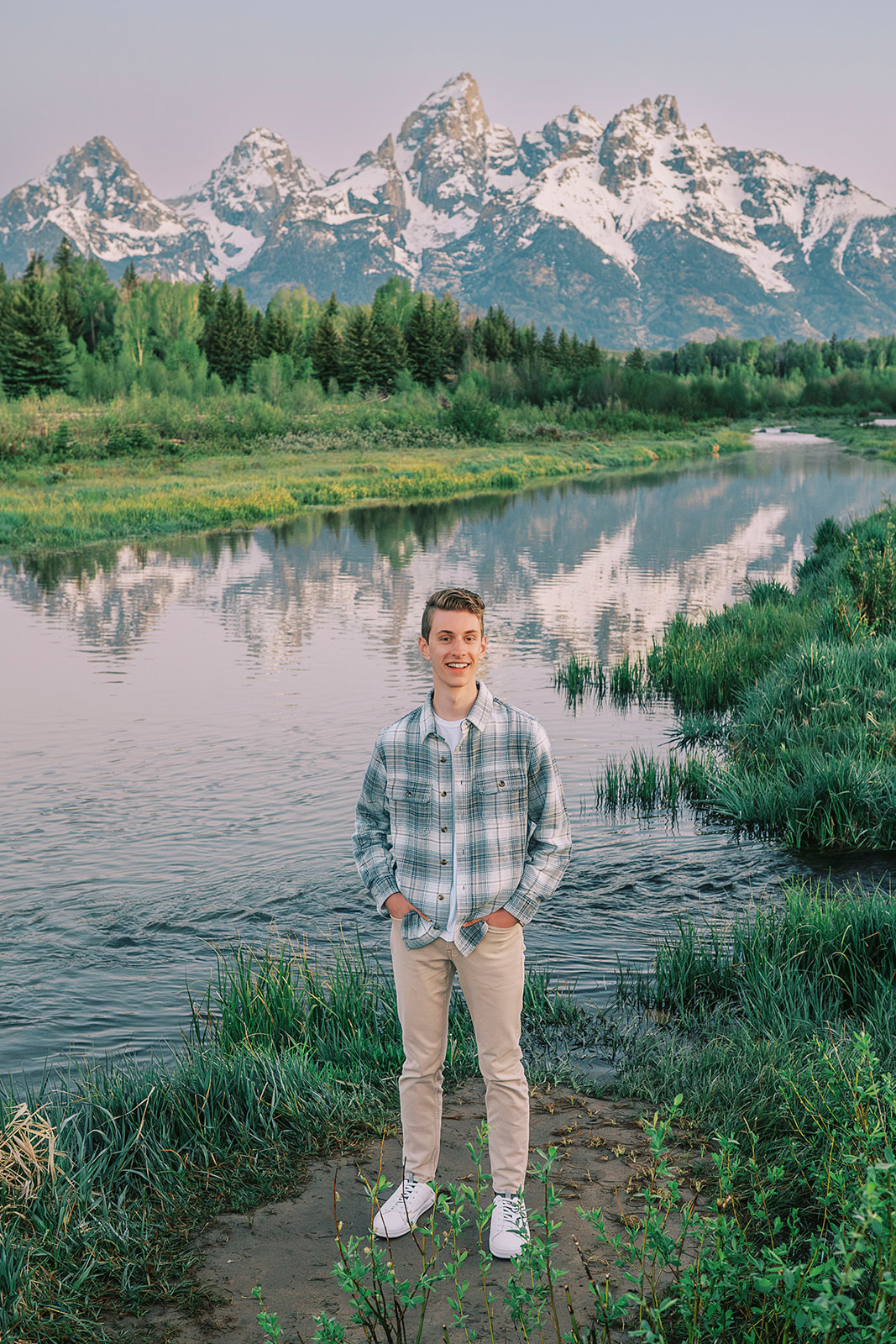 A teenager in a plaid shirt and khakis stands with hands in pockets on a riverbank at sunset
