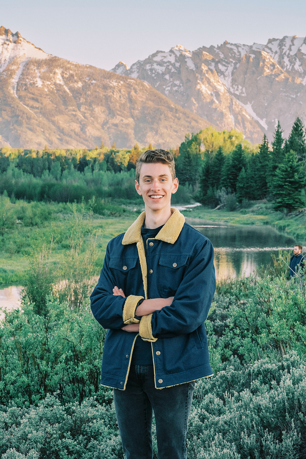 A smiling high school senior in a blue jacket and denim pants stands by a river at sunset with arms crossed after visiting colleges near Jackson Hole