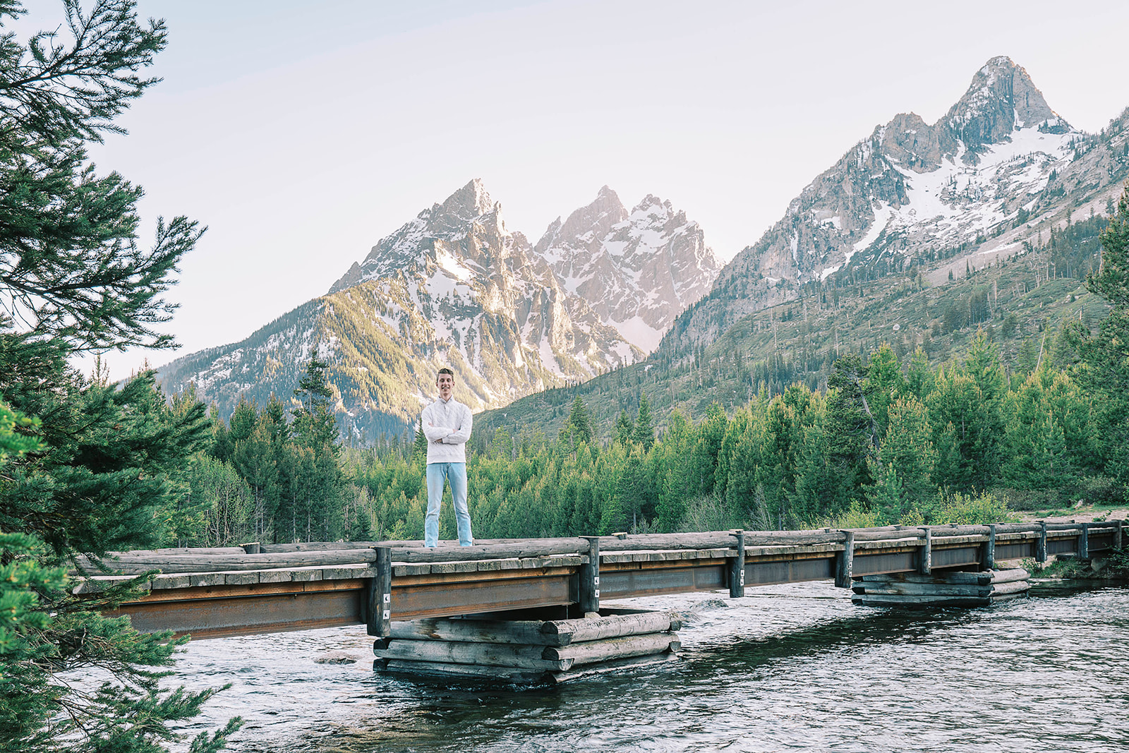 A high school senior stands on a wooden bridge over a river in the mountains with arms crossed after visiting colleges near Jackson Hole