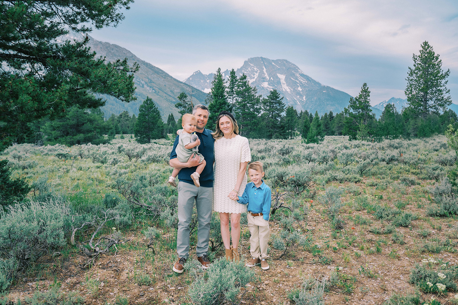 A mom and dad stand in a mountain trail holding hands with their toddler son and holding a younger child