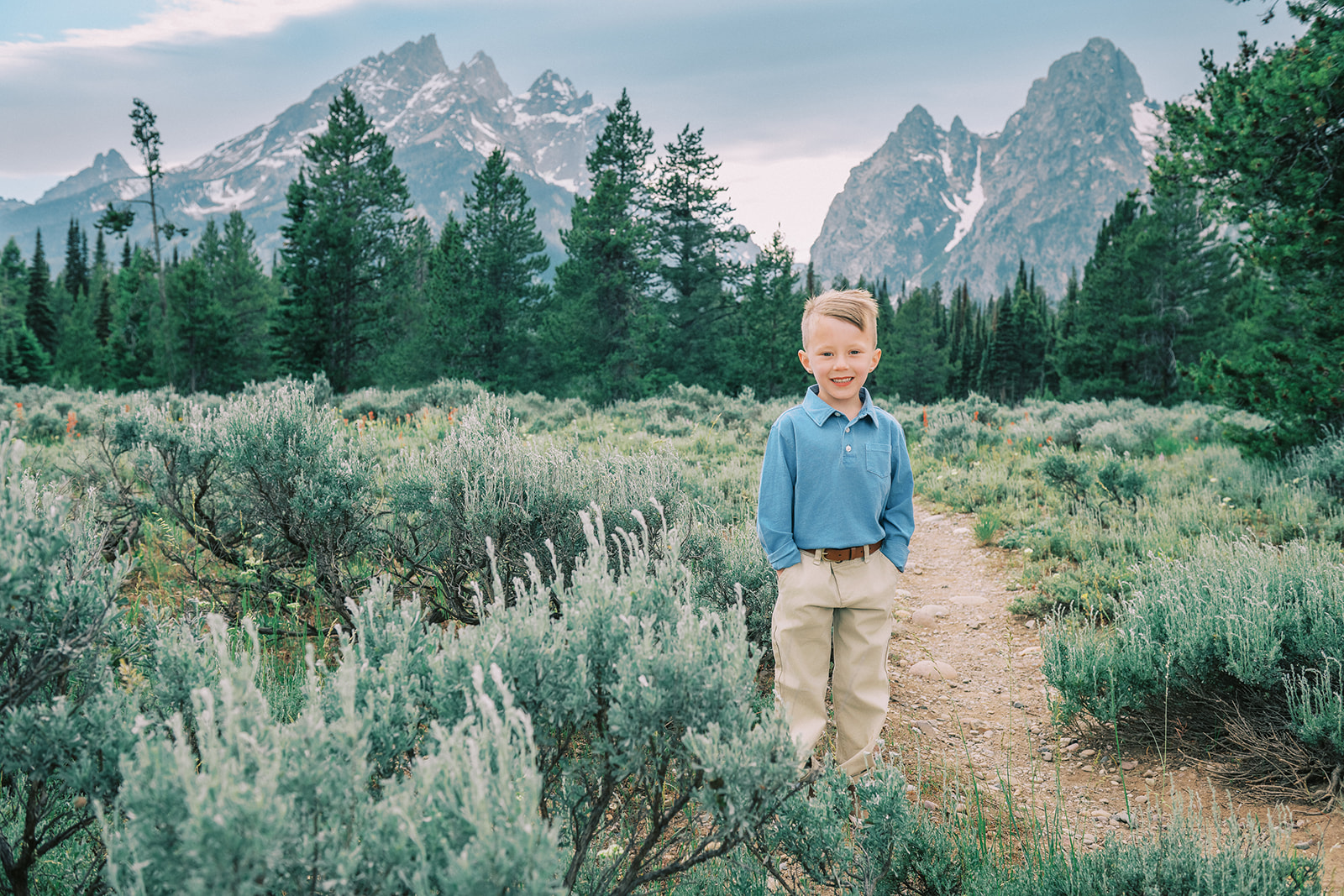 A young boy stands in a mountain trail with hands in his khaki pockets while wearing a blue shirt and smiling big