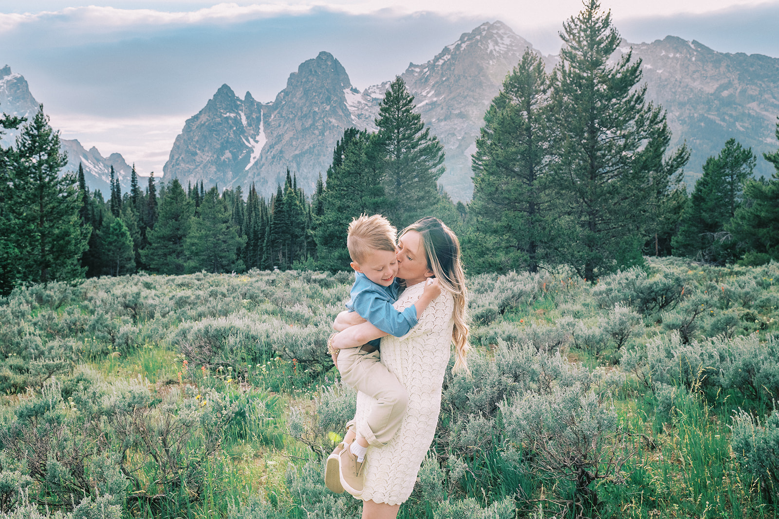 A happy mom stands in a green meadow in the mountains kissing her young son in her arms in a white dress and him in blue shirt after finding great preschools in Jackson, WY