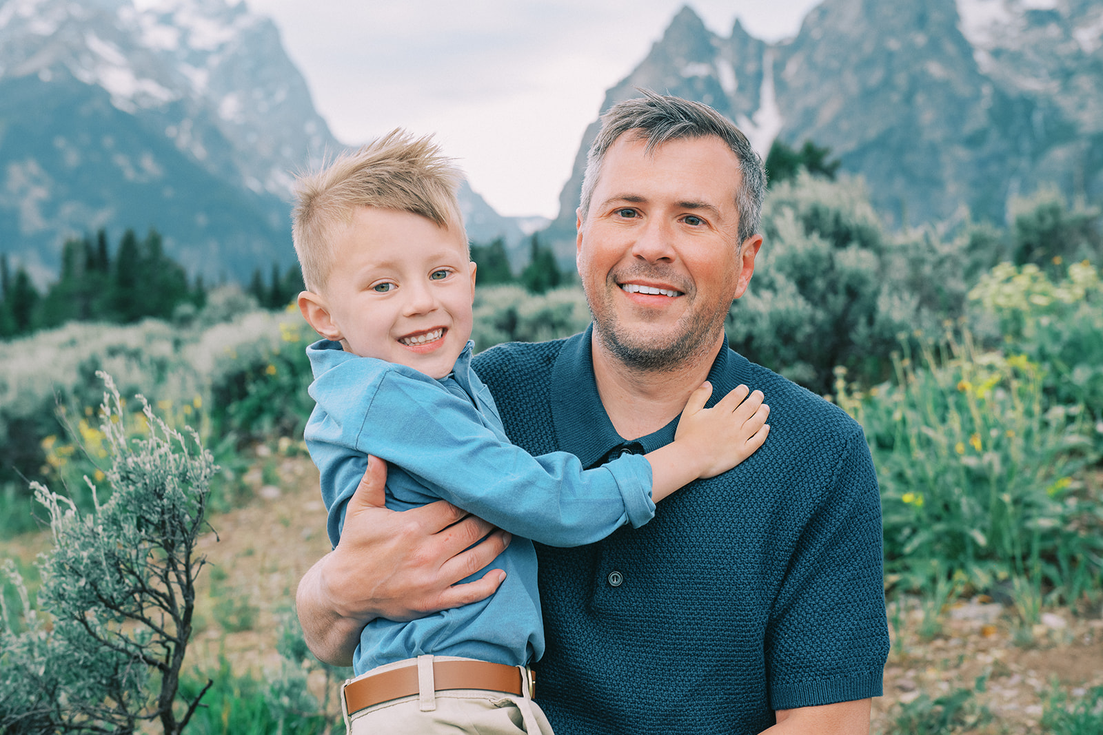 A happy dad holds his smiling young son both in blue shirts in a mountain valley after finding great preschools in Jackson, WY