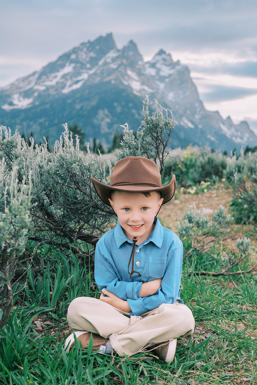 A young boy in a blue shirt sits in a meadow in a blue shirt and brown cowboy hat after attending great preschools in Jackson, WY