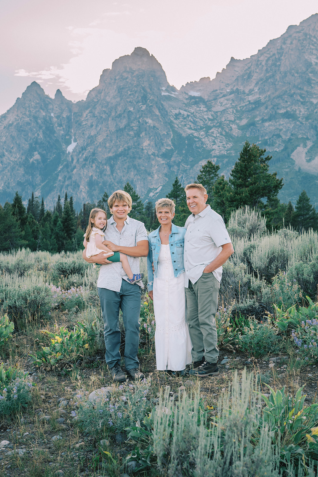 A happy family of four stand smiling in the tetons at sunset