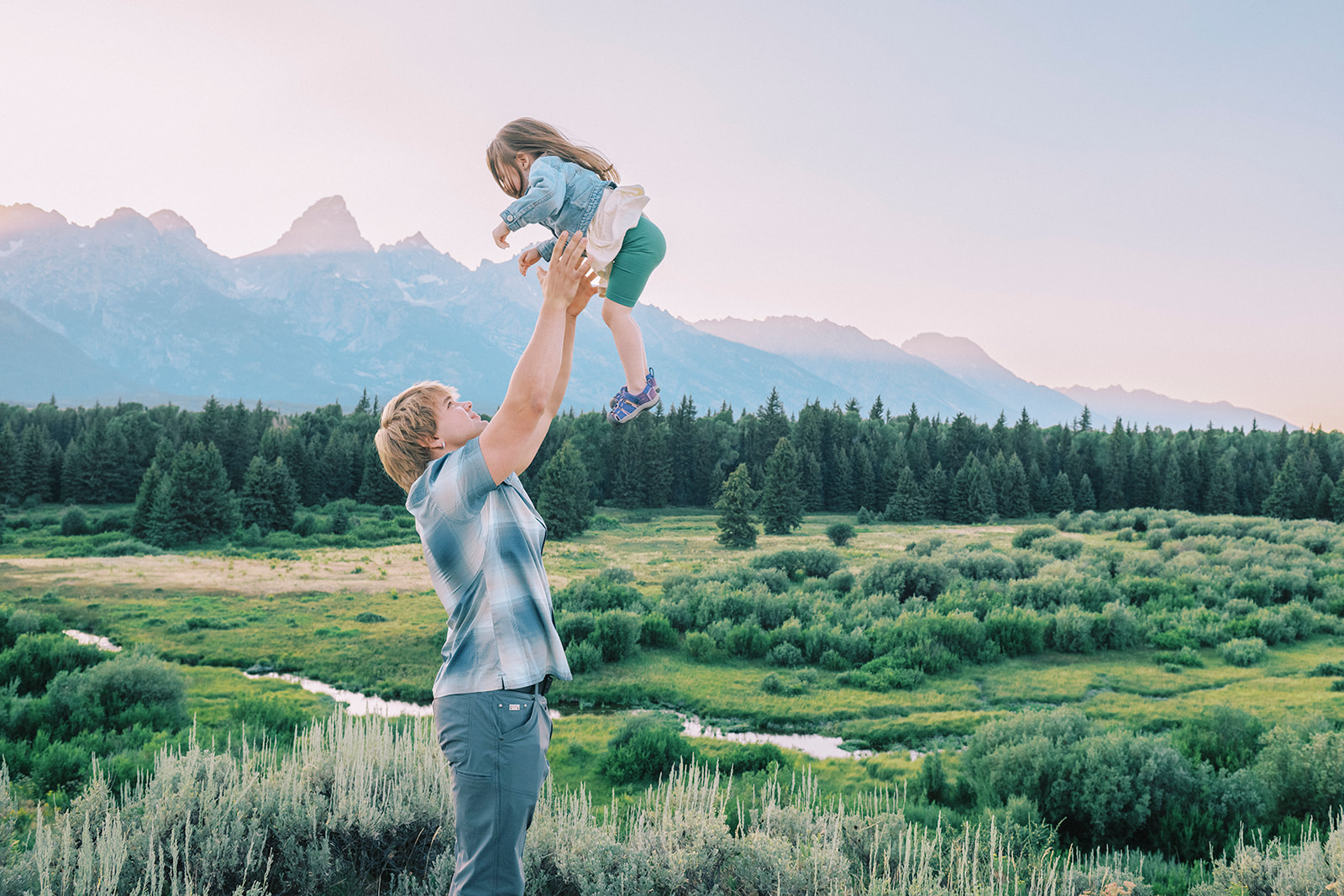 A teen in a blue b=plaid shirt tosses his younger sister in the air over his head