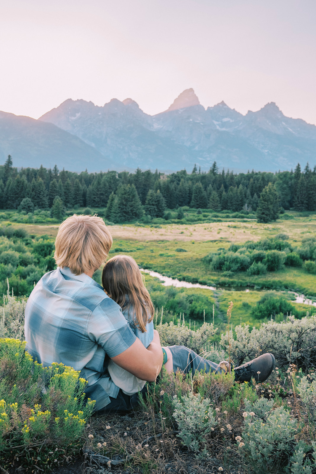 A young girl cuddles in the lap of big brother both in blue plaid while watching the sunset during activities in Grand Teton National Park