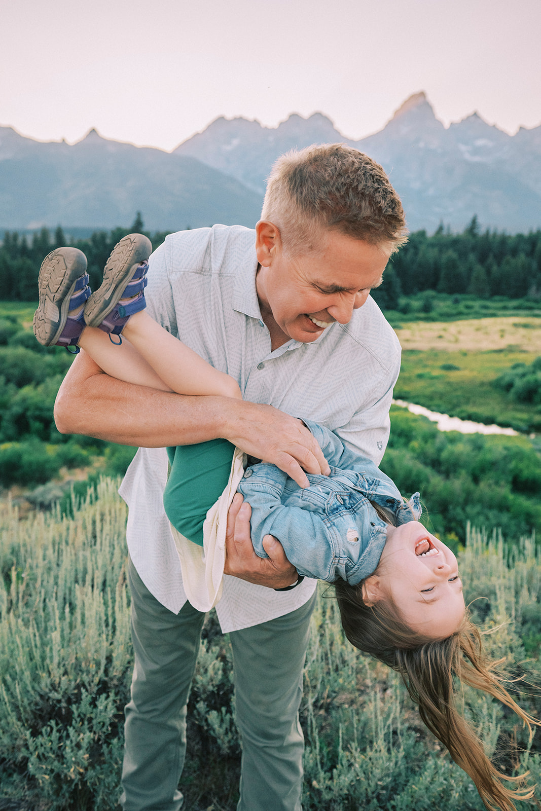 A laughing dad flips over his giggling toddler daughter while in a meadow at sunset after finding family activities in Grand Teton National Park