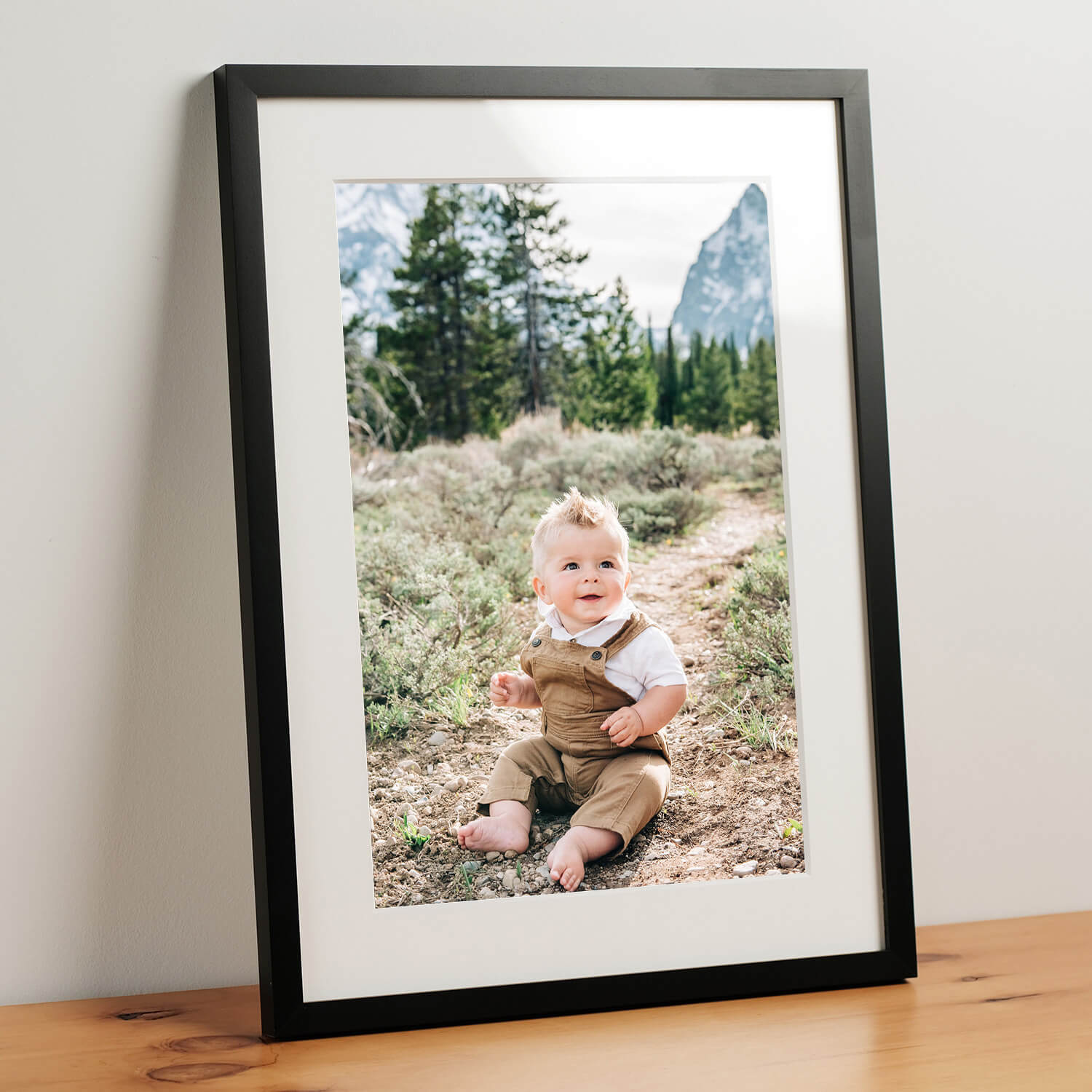 A baby portrait sitting in a mountain trail in overalls leans on a wall on a table