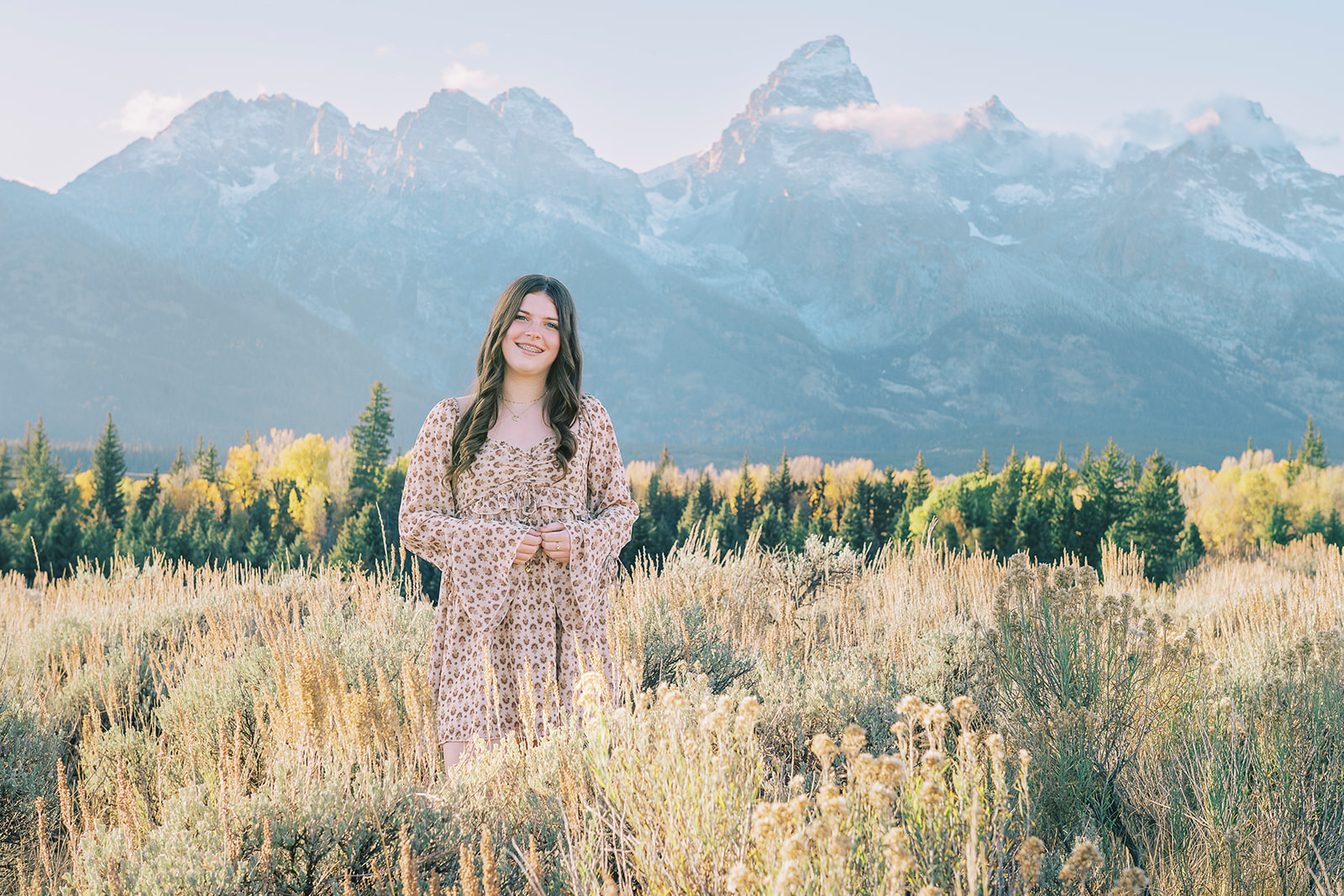 A teen girl in a red pattern dress stands in a trail through tall grasses smiling