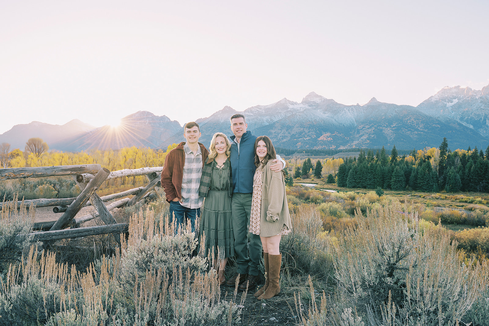 Smiling teen brother and sister stand hugging their mom and dad in a pasture at sunset while enjoying fall activities in Jackson Hole
