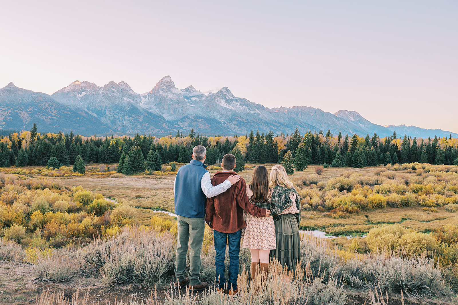 A family of four with two teens stands admiring the grand tetons in fall with hands on each other's backs while enjoying fall activities in Jackson Hole