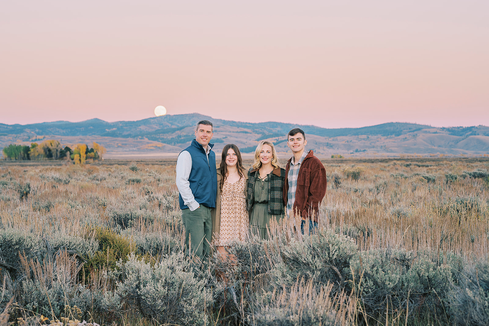 A happy mom and dad stand in a meadow at sunset with their teen son and daughter all smiling during some fall activities in Jackson Hole