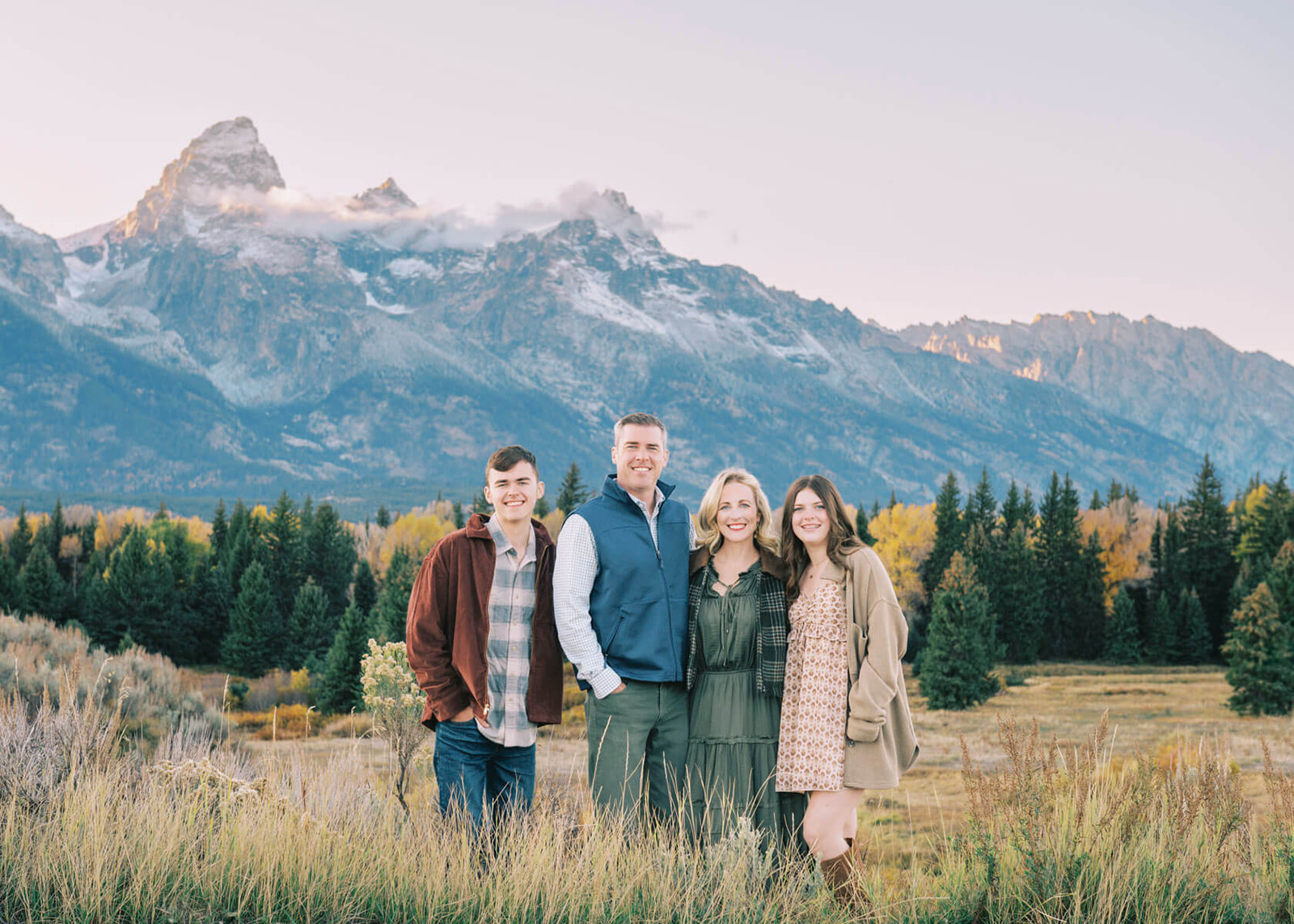 A mom and dad stand smiling with their two teen children on either side in the national park in fall