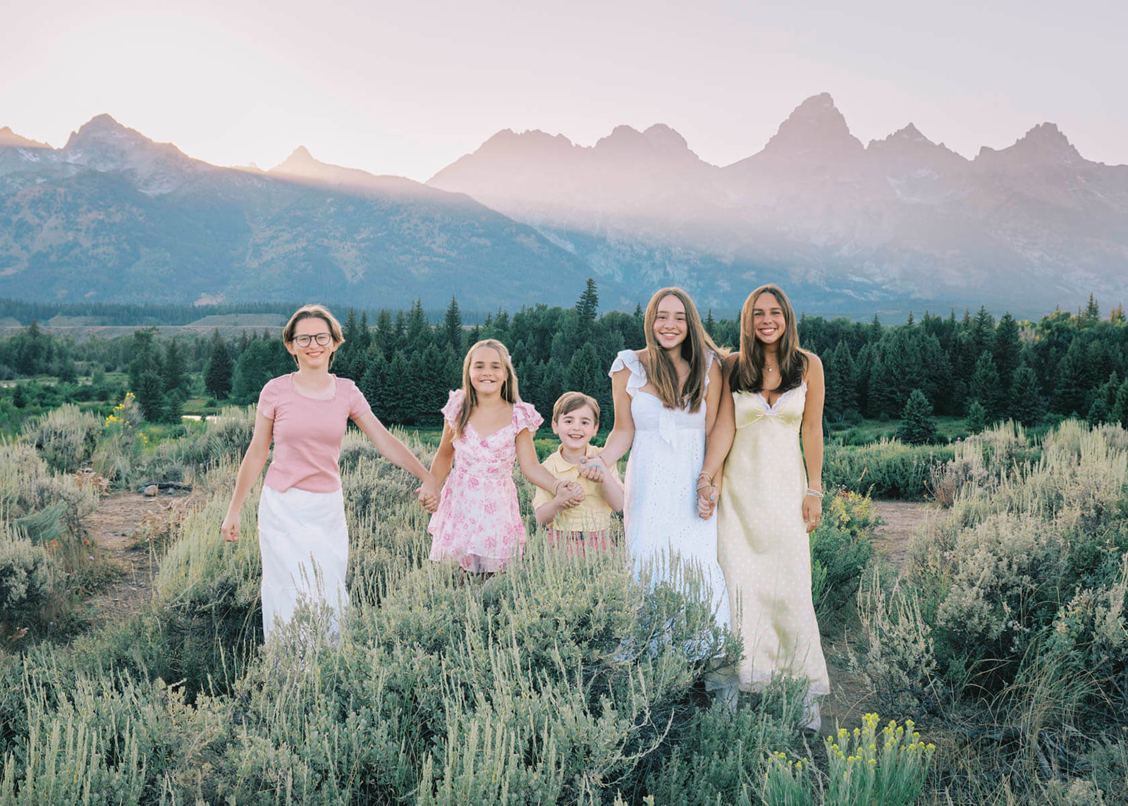 5 happy sisters in colorful dresses hold hands and smile on a hike at sunset