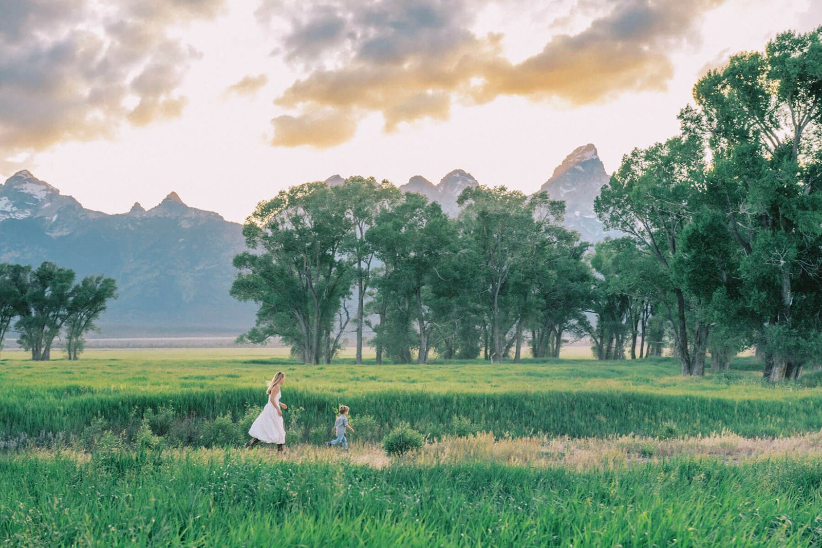 A mom in white chases after her excited toddler son running on a trail at sunset