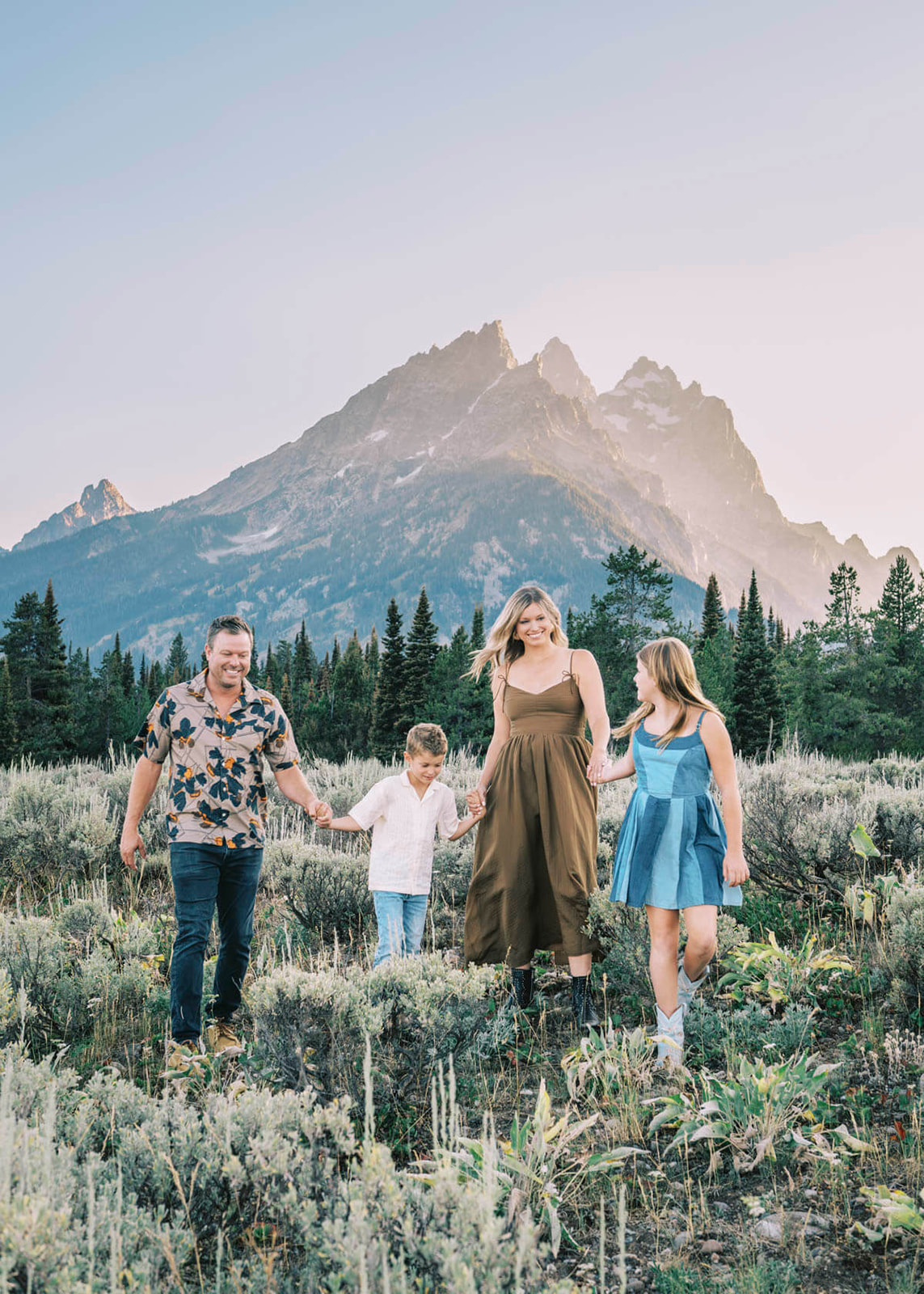 A mom and dad on a hike on a hill while holding hands with their two young children near the Grand Tetons