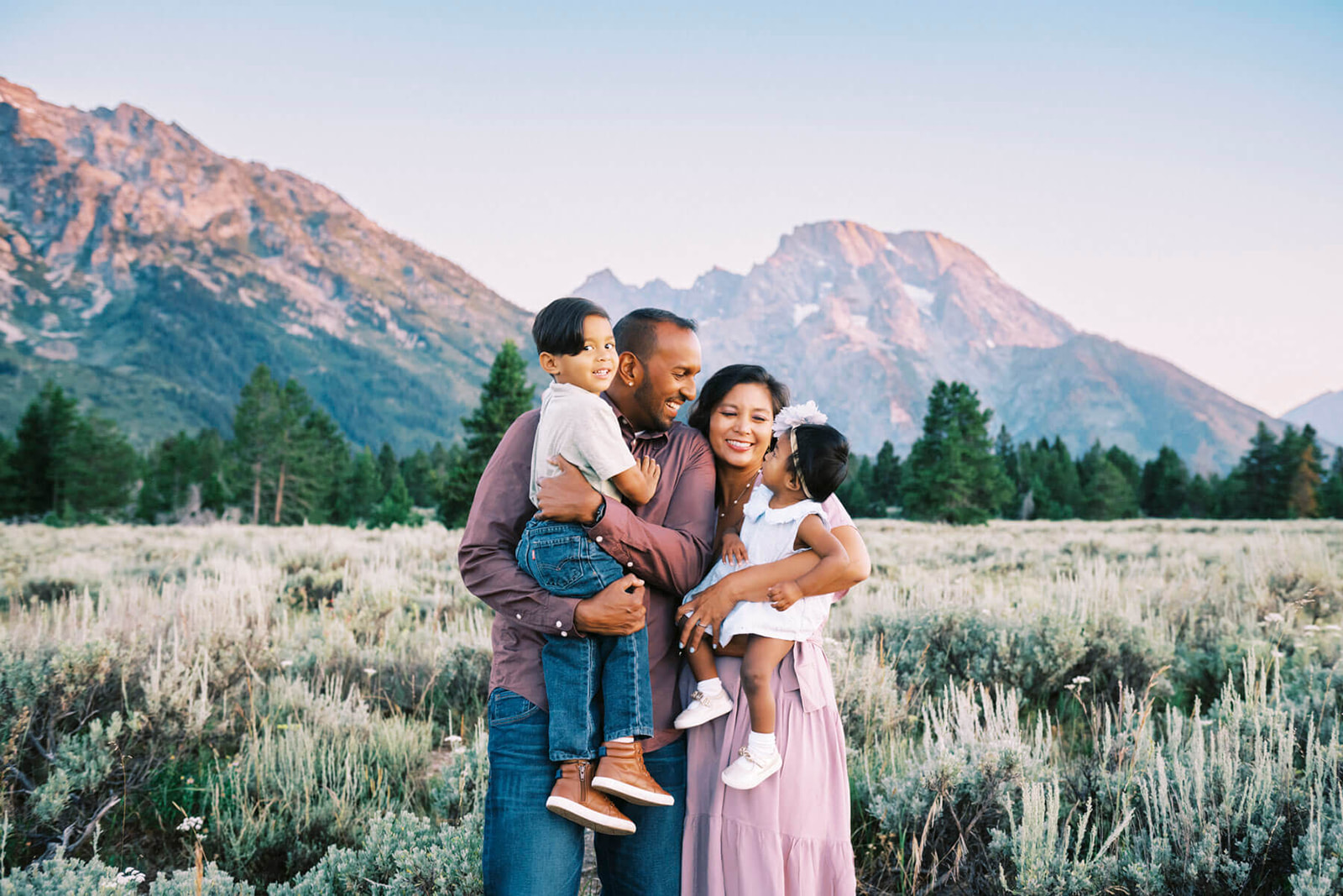 A happy family with two toddlers in mom and dad's arms laugh and stand together with the Grand Tetons behind them at sunset