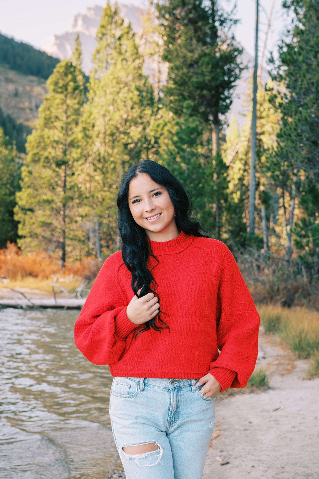 A high school senior in a red sweater and jeans plays with her hair on the edge of a river during a hike with a smile for Jenna Boshart Photography