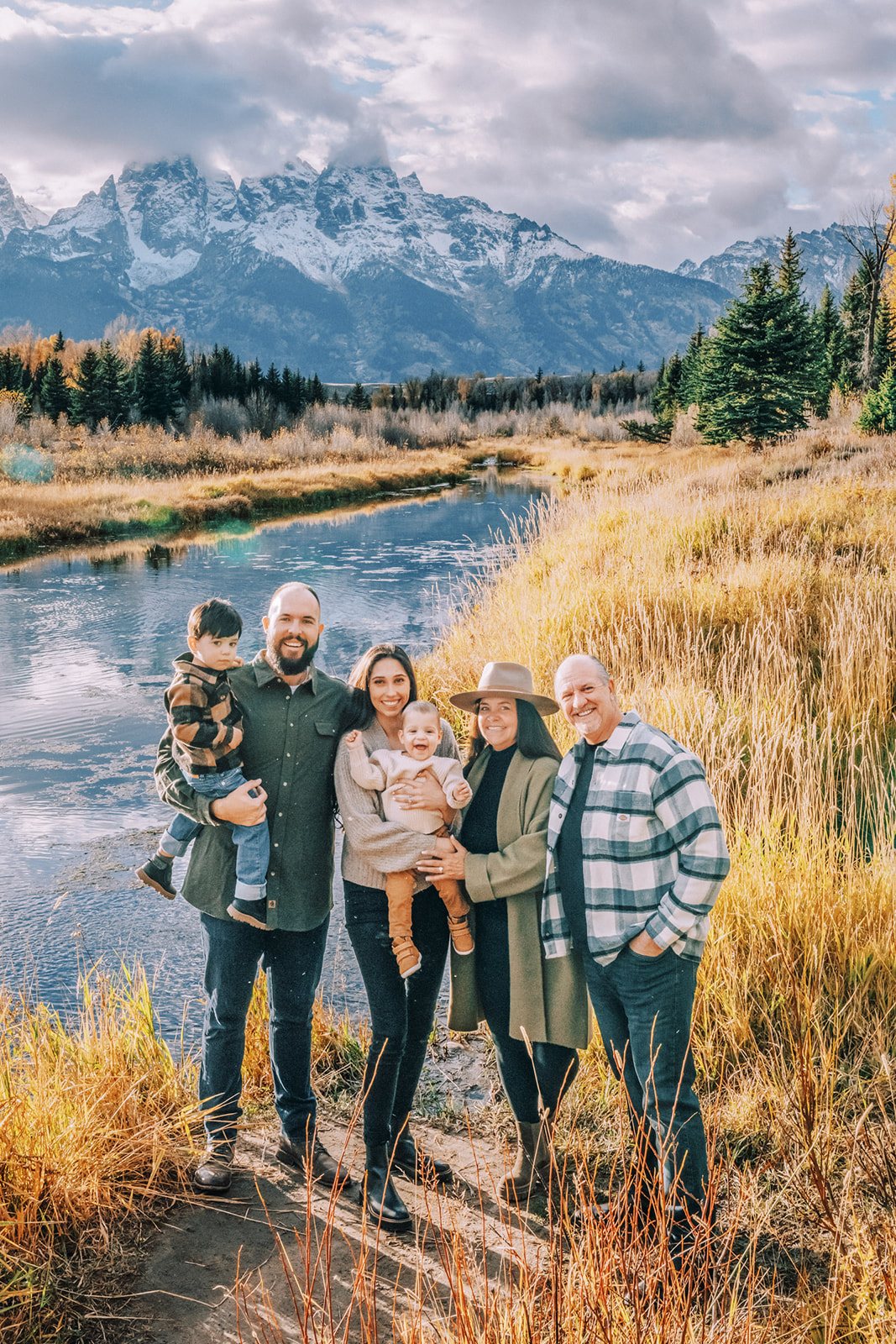A happy extended family with two toddlers stand on the edge of a river at sunset captured by Jenna Boshart Photography
