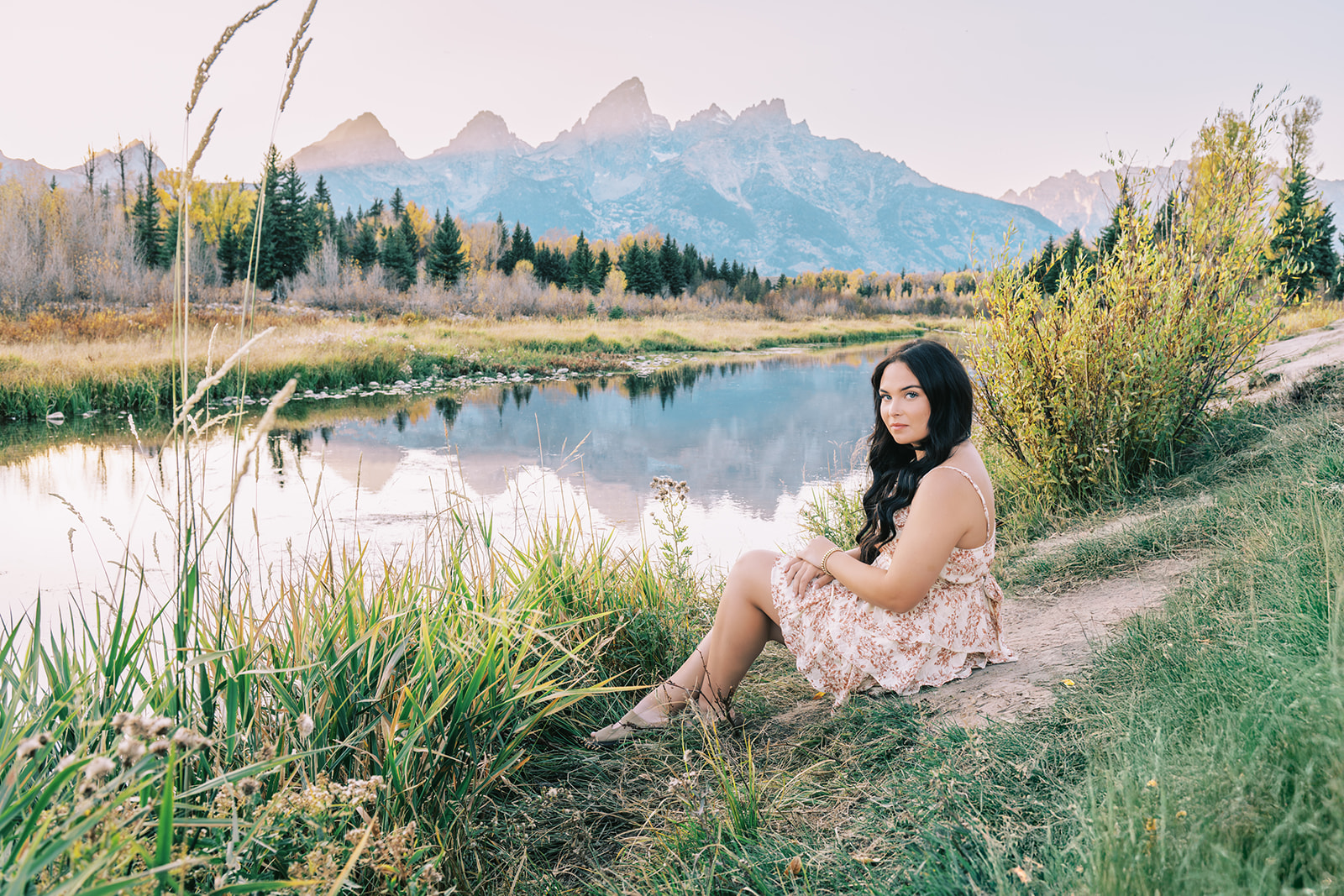 A woman sits on the edge of a river looking over her shoulder with the Tetons in the distance